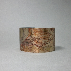 May include: A silver metal cuff bracelet with a vintage map of Natchez, Mississippi printed on it. The map is in shades of brown and gold.