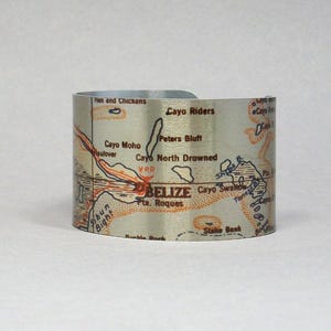 Puede incluir: Un brazalete plateado con un mapa de Belice. El mapa incluye nombres de lugares como Cayo Riders y Cayo North Drowned, con detalles en rojo y naranja. El brazalete es curvo y se encuentra sobre una superficie blanca.