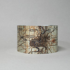 May include: A silver metal cuff bracelet with a vintage map of Bend, Oregon printed on it. The map shows the city's streets, landmarks, and surrounding areas. The map is in shades of brown, tan, and white.