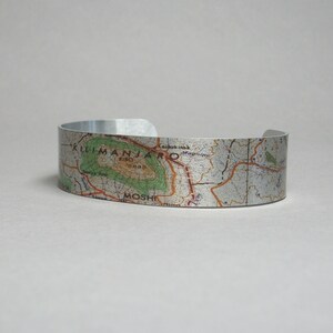 May include: A silver metal bracelet with a map of Mount Kilimanjaro printed on it. The map shows the location of Moshi, Tanzania.