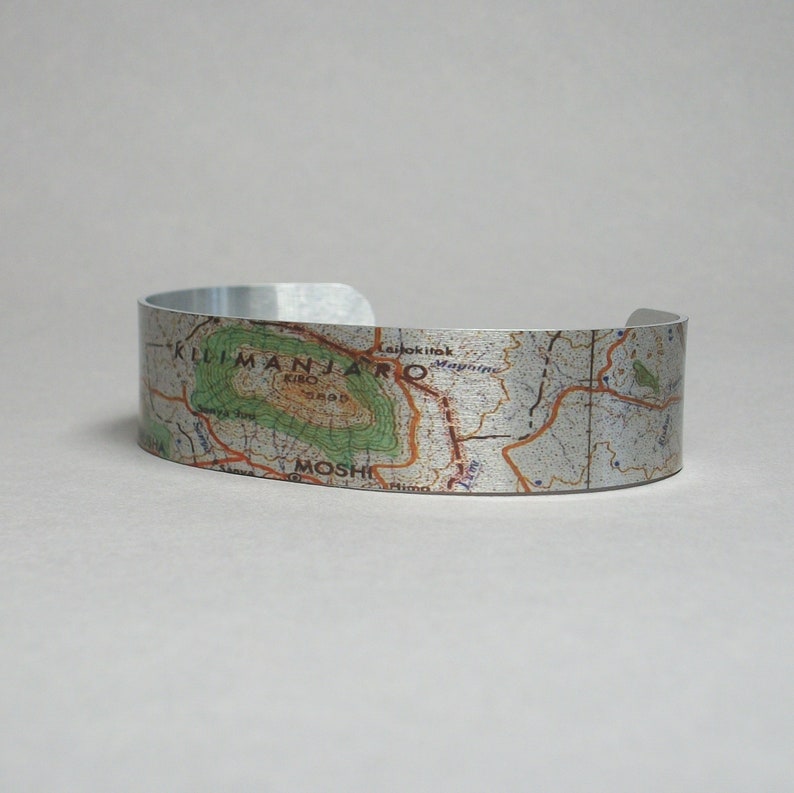 May include: A silver metal bracelet with a map of Mount Kilimanjaro printed on it. The map shows the location of Moshi, Tanzania.