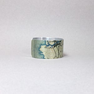May include: A silver-toned metal cuff bracelet featuring a map design in shades of blue, beige, and brown. The map details include coastlines, waterways, and topographical lines. The bracelet has a smooth, polished finish.