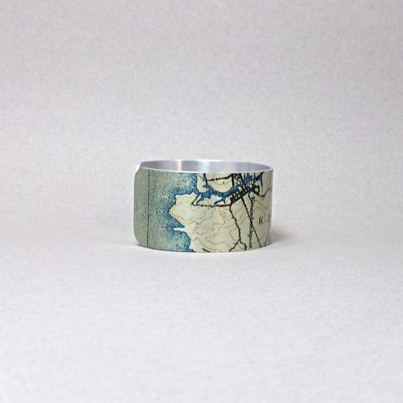 May include: A silver-toned metal cuff bracelet featuring a map design in shades of blue, beige, and brown. The map details include coastlines, waterways, and topographical lines. The bracelet has a smooth, polished finish.