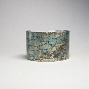 May include: A silver-toned cuff bracelet featuring a vintage map design. The map includes place names like Shagawa, Lambs, and Crossman Pr, with blue and tan hues. The bracelet has a cylindrical shape and a smooth, polished finish.