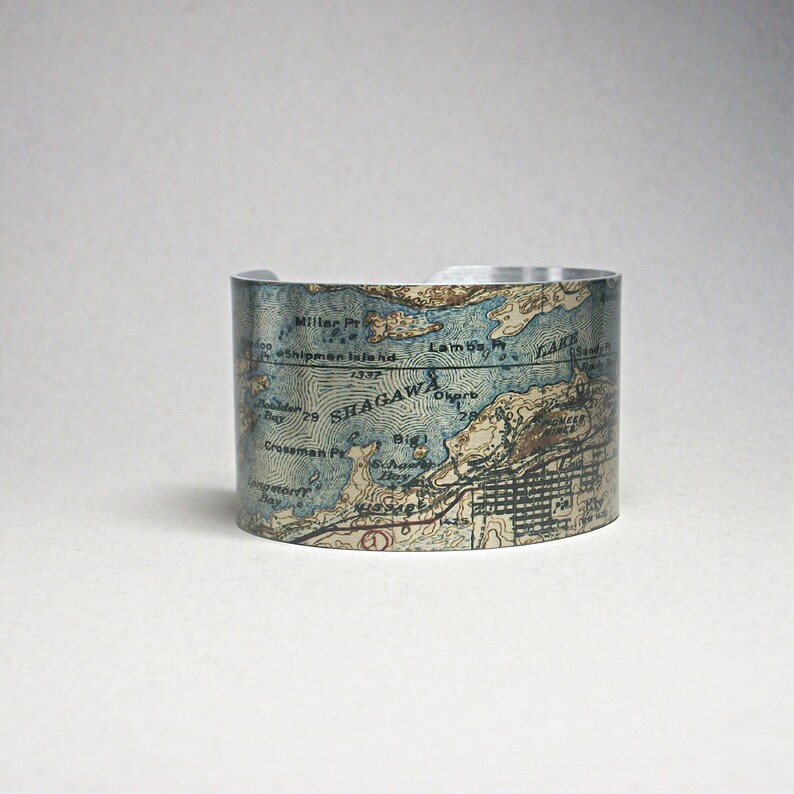 May include: A silver-toned cuff bracelet featuring a vintage map design. The map includes place names like Shagawa, Lambs, and Crossman Pr, with blue and tan hues. The bracelet has a cylindrical shape and a smooth, polished finish.