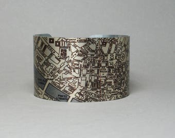 May include: A wide cuff bracelet with a map design in brown and beige tones. The map depicts a city layout, with street names and building outlines. The bracelet has a polished silver finish and is approximately 5 cm wide.