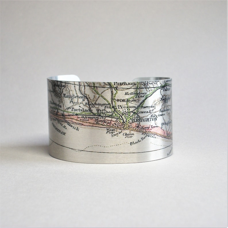 May include: A silver metal cuff bracelet featuring a vintage map of Brighton, England printed on it. The map shows the location of Brighton, Portslade, and surrounding areas. The map is in shades of green, brown, and pink.