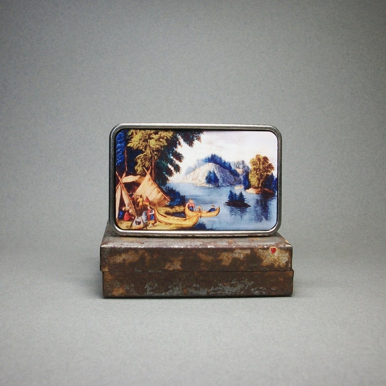 May include: A silver belt buckle with a colorful image of a lake scene with people in canoes. The image is surrounded by a silver frame.
