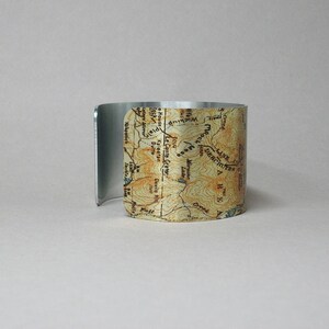 Puede incluir: Un brazalete de metal plateado con un mapa impreso de una regi&oacute;n monta&ntilde;osa. El mapa incluye los nombres de varios lugares, como "Le Conte Crater", "Wickiup", "Rock Mesa" y "Moruine Lake".
