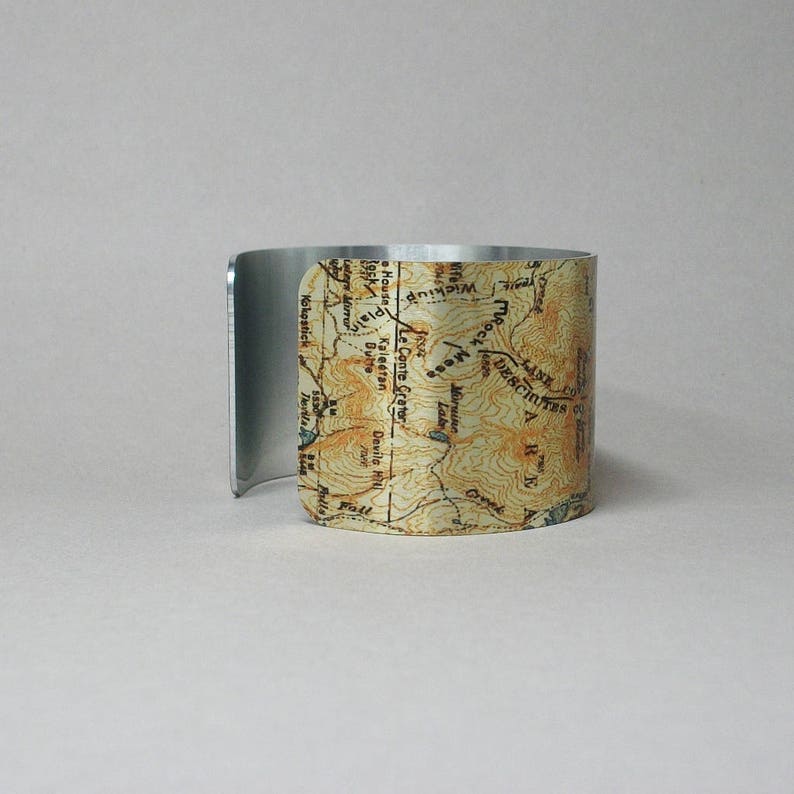 Puede incluir: Un brazalete de metal plateado con un mapa impreso de una regi&oacute;n monta&ntilde;osa. El mapa incluye los nombres de varios lugares, como "Le Conte Crater", "Wickiup", "Rock Mesa" y "Moruine Lake".