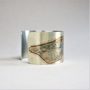 May include: A silver cuff bracelet with a vintage map of Manhattan printed on the front. The map is in shades of brown, green, and gold.