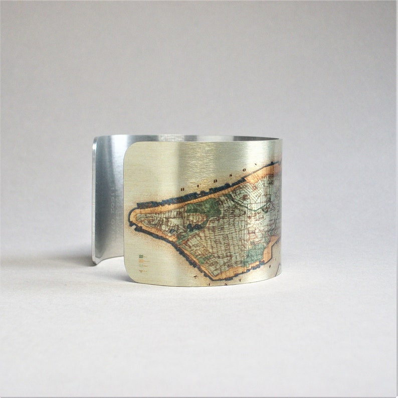 May include: A silver cuff bracelet with a vintage map of Manhattan printed on the front. The map is in shades of brown, green, and gold.