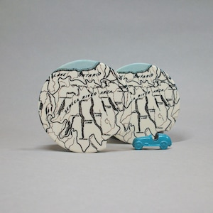 May include: Two off-white car coasters with a map design, featuring the words "Lake Ontario" and "Seneca River Area". A small, turquoise toy car sits in front of the coasters. The coasters have a light blue trim.