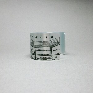 May include: A silver-toned metal cuff bracelet featuring a black printed map design. The map includes text labels such as Mendota, City Bath House, Sherman St, Johnson St, Blair St, Washington St, Brearly St, Railroad St, Wilson St, and State Capitol.