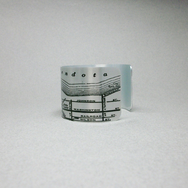 May include: A silver-toned metal cuff bracelet featuring a black printed map design. The map includes text labels such as Mendota, City Bath House, Sherman St, Johnson St, Blair St, Washington St, Brearly St, Railroad St, Wilson St, and State Capitol.
