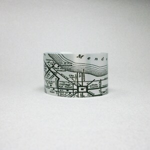 May include: A silver-toned metal cuff bracelet featuring a black printed map of Madison, Wisconsin. The design displays street names like State Street, landmarks such as the State Capitol and the University of Wisconsin Boathouse, and Lake Mendota.