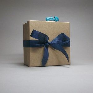 May include: A square, light brown cardboard gift box wrapped with a dark blue ribbon tied in a bow. A small, bright turquoise toy car is positioned on the top center edge of the box, resting horizontally against the lid's top surface.