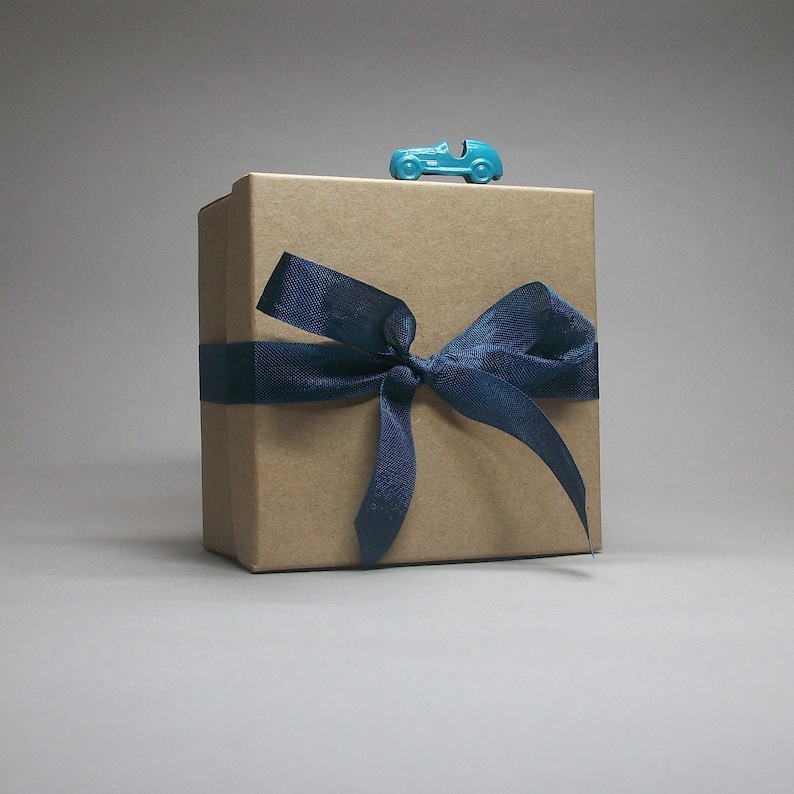 May include: A square, light brown cardboard gift box wrapped with a dark blue ribbon tied in a bow. A small, bright turquoise toy car is positioned on the top center edge of the box, resting horizontally against the lid's top surface.