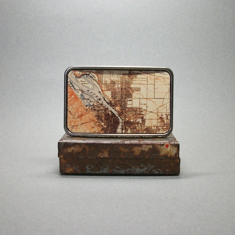 May include: A silver belt buckle with a map of a city on it. The map is in shades of brown and white and shows a river running through the city. The buckle is sitting on a rusty metal box.