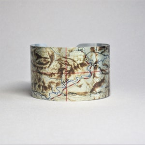 May include: A silver metal cuff bracelet with a topographic map printed on it. The map shows a river, mountains, and the words "Hoh" and "Nolan".