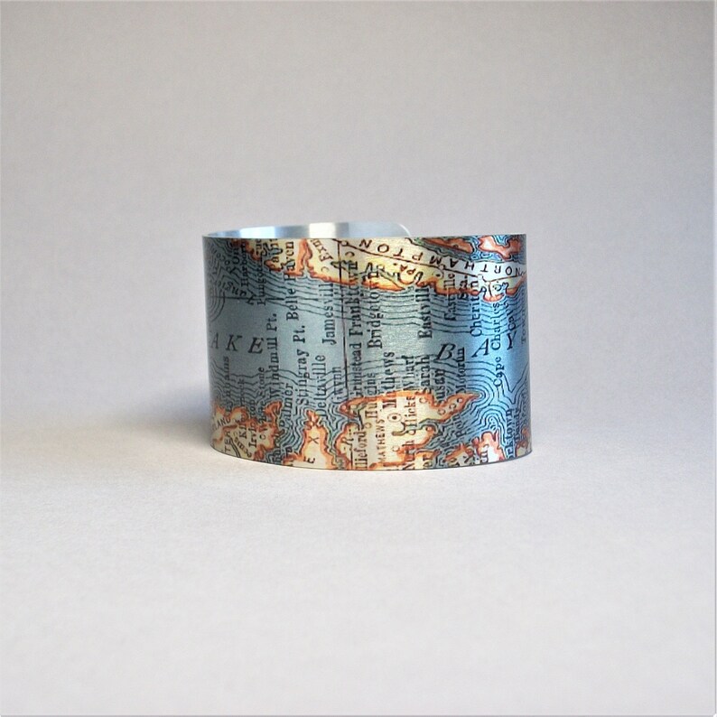 May include: A silver cuff bracelet with a vintage map design. The map features blue water, brown land, and white text. The text on the map includes "AKE", "Stingray Pt. Belle Haven", "Jamesville", "Mathews", "Eastville", "Cape Charles", and "NORTHAMPTON".