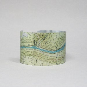 May include: A silver metal cuff bracelet with a topographic map design. The map features a river, contour lines, and the text "RIVER".