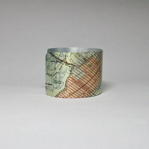 May include: A silver metal cuff bracelet with a vintage map design. The map is printed on the cuff and shows a city with streets and waterways. The cuff is a light beige color with brown and blue accents.
