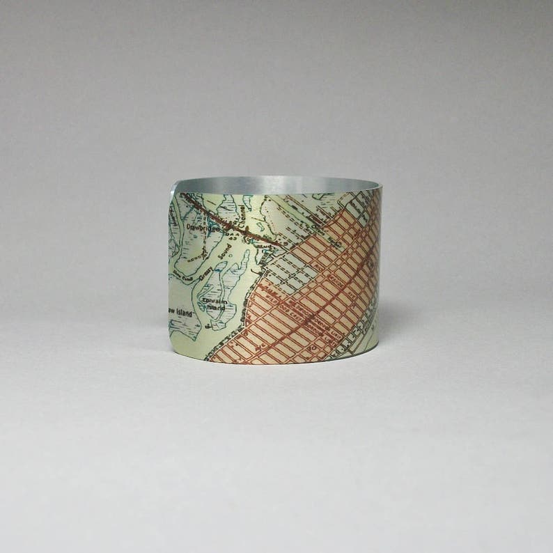 May include: A silver metal cuff bracelet with a vintage map design. The map is printed on the cuff and shows a city with streets and waterways. The cuff is a light beige color with brown and blue accents.