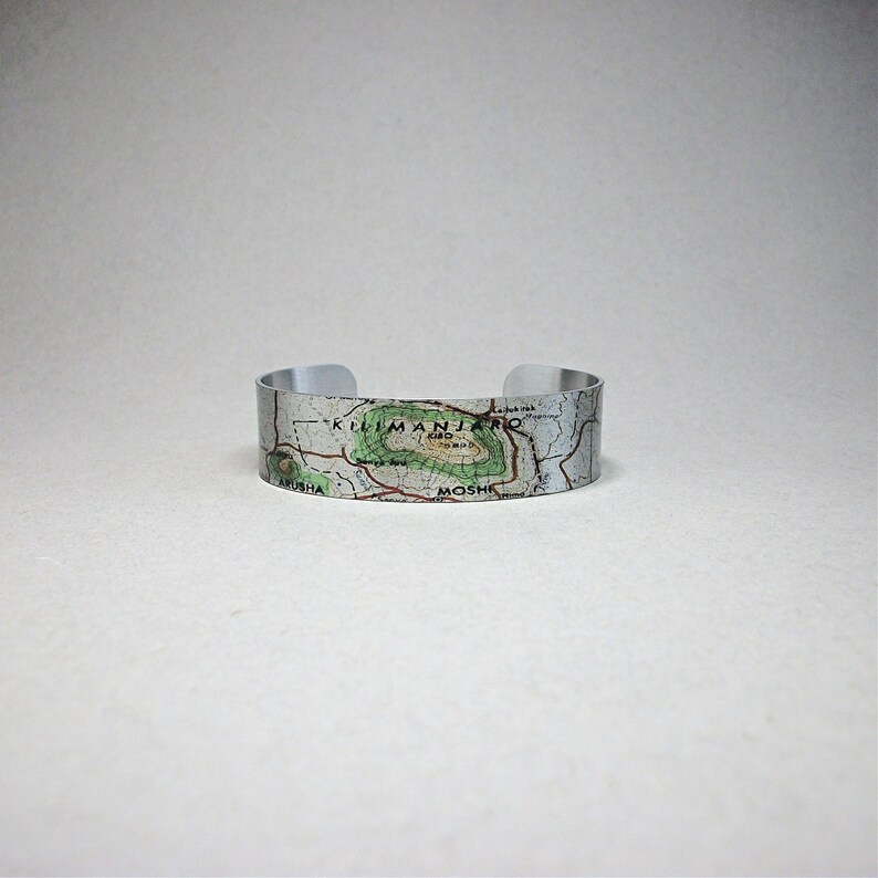 May include: A silver metal bracelet with a map of Kilimanjaro printed on it. The map shows the location of Moshi and Arusha. The bracelet is on a white surface.