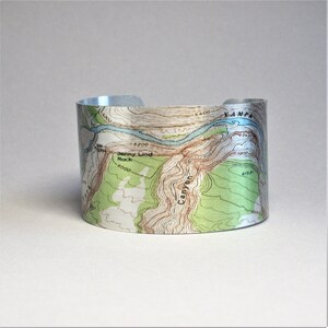 May include: A silver metal cuff bracelet with a topographic map design. The map shows a river, mountains, and the text "Jenny Lind Rock" and "Yampa Canyon".
