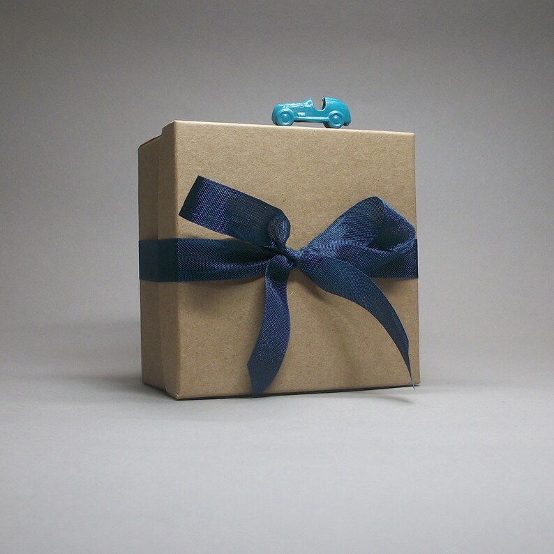 May include: A kraft paper gift box tied with a navy blue ribbon and bow. A small, turquoise toy car sits on top of the box. The box is square and the background is a solid gray.