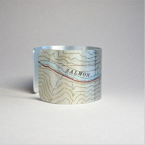 May include: A silver metal cuff bracelet with a topographic map design featuring the word "SALMON" and contour lines.