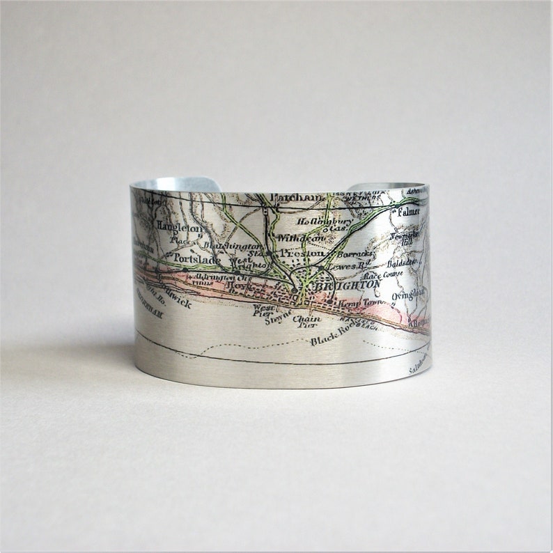 May include: A silver metal cuff bracelet featuring a vintage map of Brighton, England printed on it. The map shows the coastline and surrounding areas, including the names of streets and landmarks.