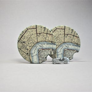 May include: Two round coasters with a map of New Orleans printed on them. The coasters are beige with blue accents and the text "NEW ORLEANS" is printed on each coaster. A small silver toy car is sitting in front of the coasters.