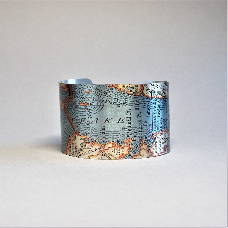 May include: A silver metal cuff bracelet with a vintage map design. The map is printed on the metal and shows a coastline with the words "PEAKE" and "UNDERLAND" visible.