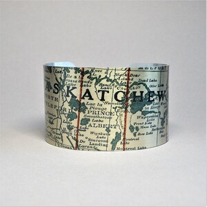 May include: A silver-colored cuff bracelet featuring a vintage map of Saskatchewan, Canada. The map includes place names like Prince Albert and Churchill, with red vertical lines and a light blue interior.