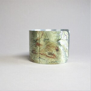 Pu&ograve; includere: Un braccialetto rigido in metallo argentato con un design a mappa topografica. La mappa presenta linee e testo in marrone, verde e bianco. Il testo sulla mappa include "Greene Village" e "Arnold Brook".