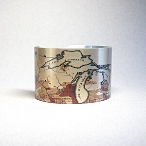 Peut inclure: Un bracelet manchette argenté orné d'une carte vintage. La carte représente la région des Grands Lacs, dont le lac Supérieur et le lac Michigan, avec un fond sépia et des accents rouges. Le bracelet est large et ouvert.