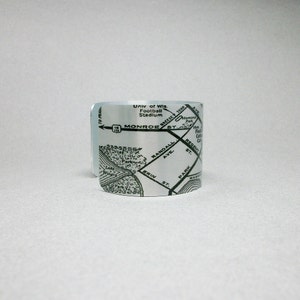 May include: A silver-toned metal cuff bracelet featuring a black printed map design. The map displays street names including Monroe St, Randall Ave, and Erin St, along with landmarks like the Univ of Wis Football Stadium and Lake Wingra.