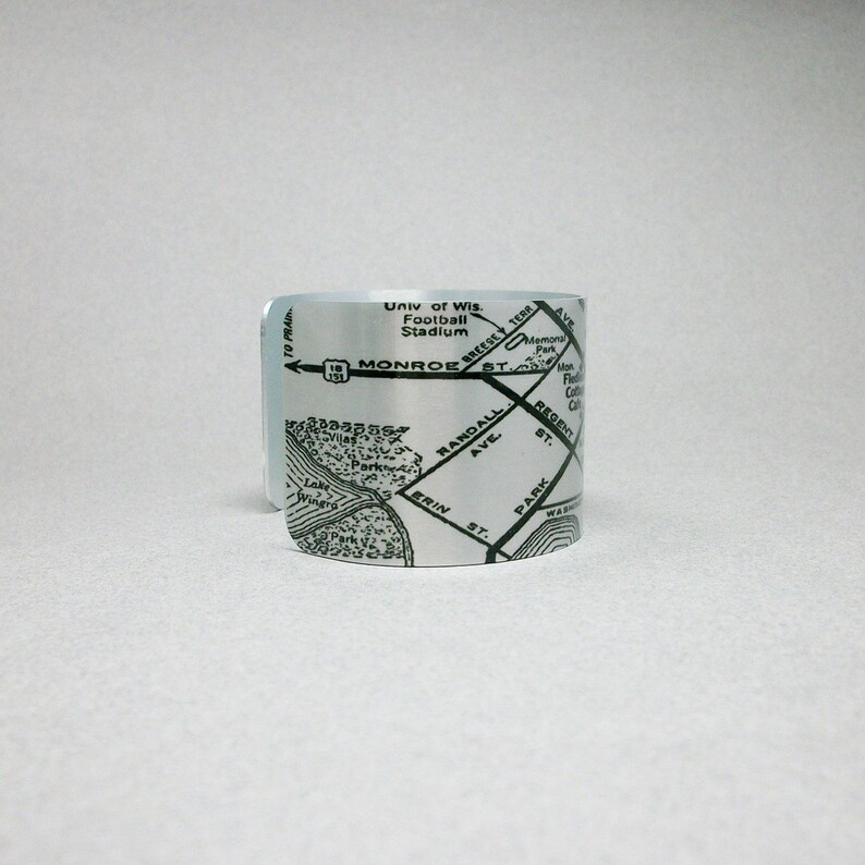 May include: A silver-toned metal cuff bracelet featuring a black printed map design. The map displays street names including Monroe St, Randall Ave, and Erin St, along with landmarks like the Univ of Wis Football Stadium and Lake Wingra.