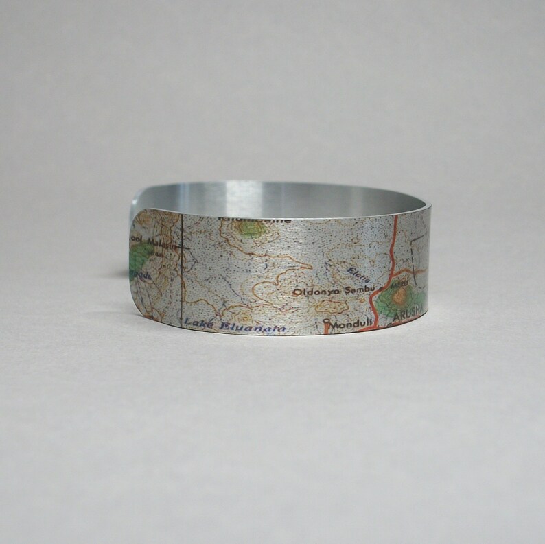 May include: A silver metal bracelet with a map of a region in Tanzania printed on it. The map shows the names of locations like Lake Manyara, Oldonyo Sambu, and Mondul.