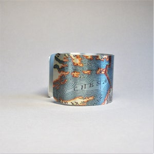 May include: A silver cuff bracelet with a map of the Chesapeake Bay printed on it. The map is in shades of blue, green, and orange.