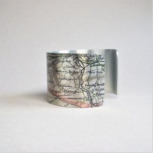 May include: A silver metal cuff bracelet featuring a vintage map design. The map shows the names of towns and rivers in black lettering. The map is printed on a white background with green and brown lines.