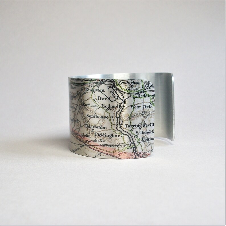 May include: A silver metal cuff bracelet featuring a vintage map design. The map shows the names of towns and rivers in black lettering. The map is printed on a white background with green and brown lines.