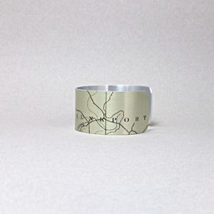 May include: A silver-toned metal cuff bracelet with a map design. The map features black lines and text, with a beige and green background. The word "PORT" is visible on the bracelet.