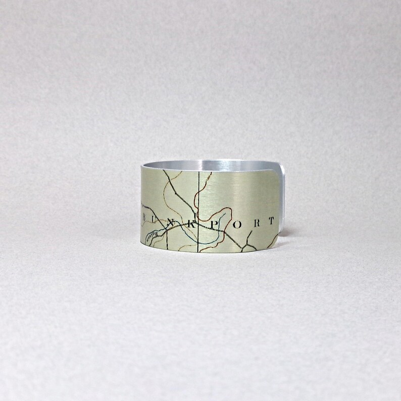 May include: A silver-toned metal cuff bracelet with a map design. The map features black lines and text, with a beige and green background. The word "PORT" is visible on the bracelet.