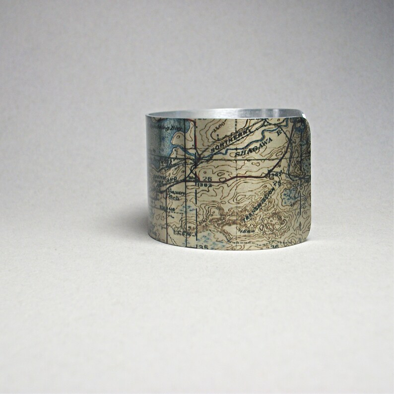 May include: A silver cuff bracelet with a map design. The bracelet features a detailed topographic map print in shades of tan, blue, and brown. The map design wraps around the entire bracelet, creating a unique and stylish accessory.