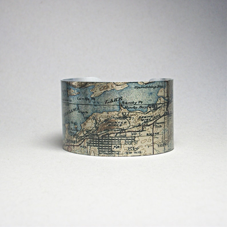 May include: A silver cuff bracelet featuring a vintage map design. The map includes blue water features and brown landmasses, with place names visible. The bracelet has a curved shape and is displayed against a white background.