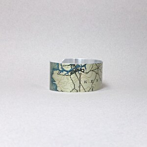 May include: A silver cuff bracelet with a map design. The map features blue and tan tones, with the word "KEN" visible. The bracelet is open at the bottom and has a smooth, metallic finish.