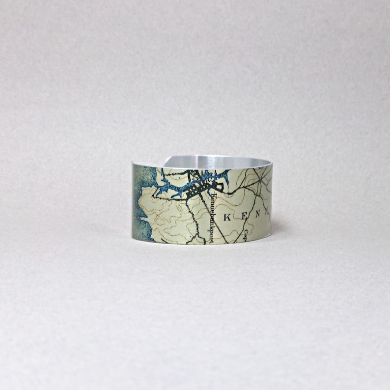 May include: A silver cuff bracelet with a map design. The map features blue and tan tones, with the word "KEN" visible. The bracelet is open at the bottom and has a smooth, metallic finish.